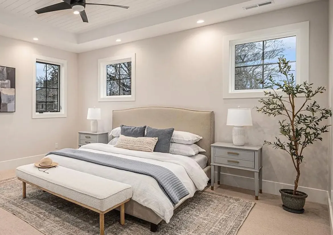 Neutral bedroom with an upholstered bed, gray nightstands, a bench, and a potted olive tree.