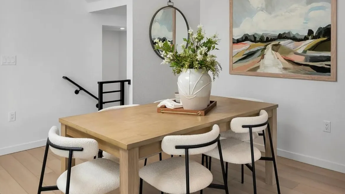 Bright dining room with a light wood table, white boucle chairs on black legs, a white vase of greenery on a wooden tray, a strap-hung round mirror, and an earthy abstract landscape painting.