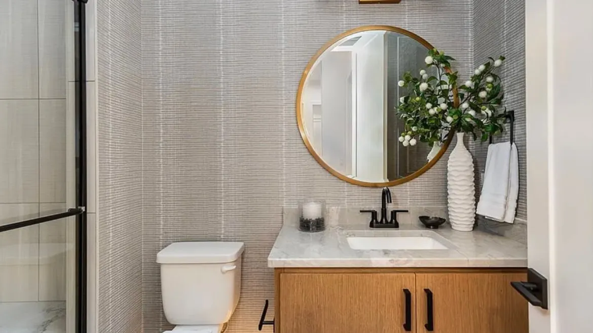 Bathroom with textured gray wallpaper, a gold-framed round mirror, a light wood vanity with marble top, matte black faucet, and a white ribbed vase with greenery.